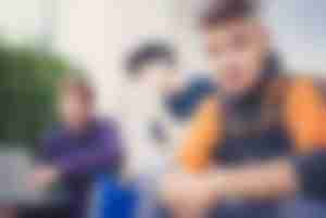 Teilnehmer der Produktionsschule: Jugendlicher mit kurzen Haaren und orange/blauer Kleidung, dahinter zwei weitere Jugendliche, die in einen Laptop schauen